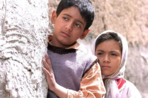 Why Victory Sometimes Feels Sad in Cinema 1 A poignant medium shot of two young Iranian children, a boy and a girl, peering anxiously from behind a textured concrete wall. Their somber, melancholic expressions contrast with traditional narratives of triumph, hinting at a victory that brings more emotional burden than joy.