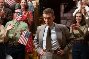 Why Victory Sometimes Feels Sad in Cinema 4 A medium shot of Cillian Murphy as J. Robert Oppenheimer standing amidst a cheering crowd waving American flags. Despite the celebratory atmosphere of a nationalist victory, his expression is deeply conflicted, hollow, and haunted, capturing the psychological weight and tragic undercurrent of his scientific triumph.