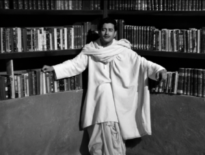 Pyaasa star Guru Dutt performing the song “Jaane Woh Kaise Log The” in a black-and-white library scene, dressed in traditional attire and leaning against curved bookshelves filled with books.